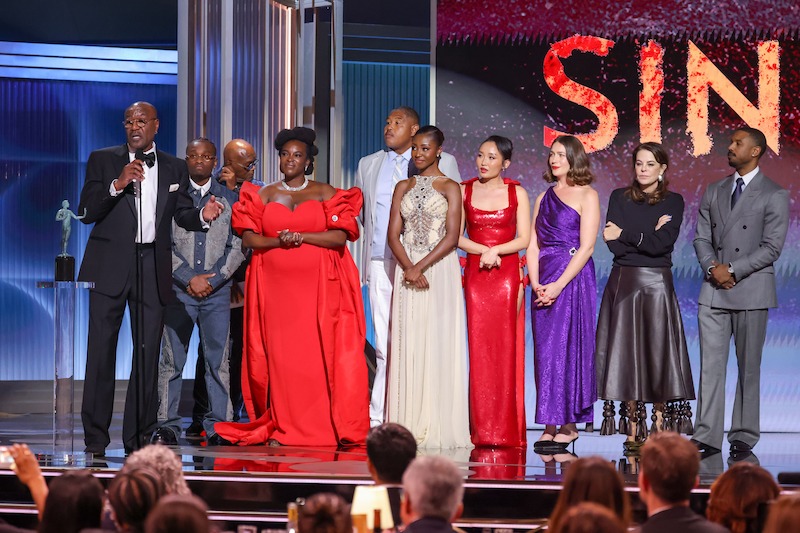 A group of well-dressed actors stands on stage in front of an audience at an awards ceremony, with microphones and bright lights, giving an acceptance speech.