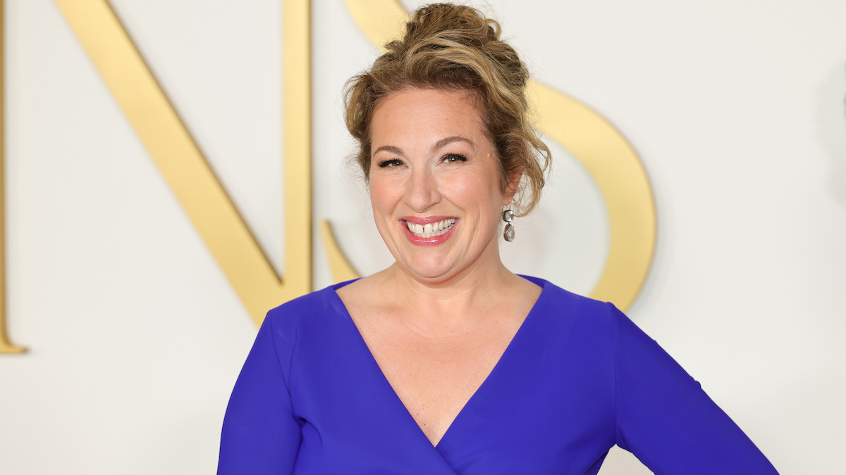 Woman with light hair in an updo, wearing a bright blue dress, smiling and posing in front of a white and gold backdrop at a formal event.