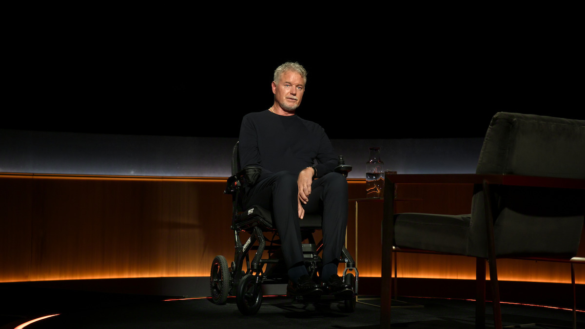 Eric Dane sits in a wheelchair on a dimly lit stage next to an empty chair and a small table with a glass carafe, in a modern studio interview setting with warm lighting and dark background.