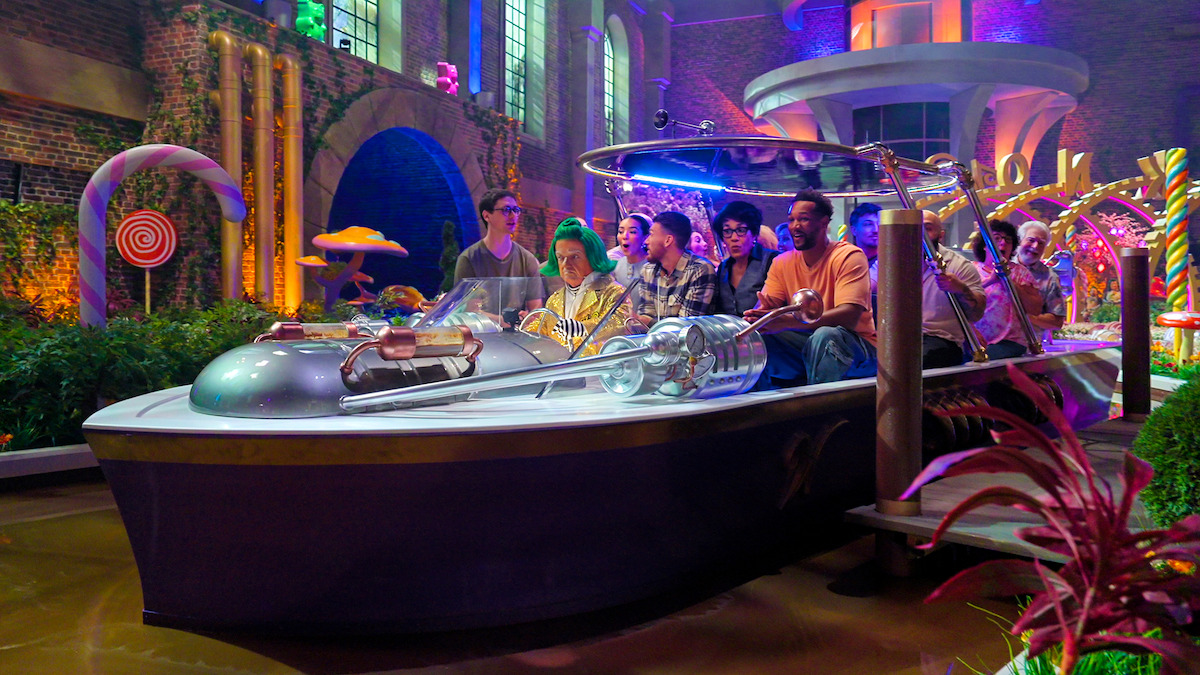 A group of people ride together in a boat through a colorful, whimsical indoor setting with candy decorations, glowing lights, lush plants, and fantasy architecture.