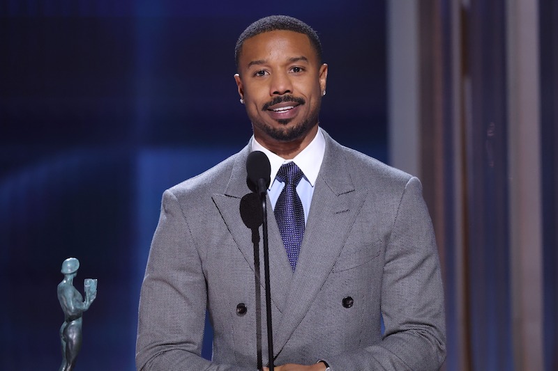 Michael B. Jordan in a gray suit speaking at a microphone on stage, with an award statue visible to the side, in a formal event or ceremony setting with blue-lit background.