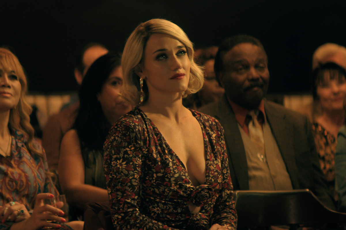 Alice Kremelberg as Anneliese Shaw, with blonde hair in floral dress sits among an audience in a dimly lit room, looking attentive. Other people are seated around her, suggesting a formal or social event.