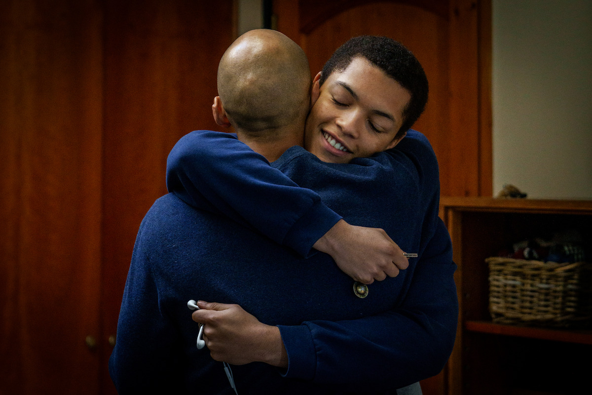 Two people hugging warmly indoors, one smiling with eyes closed. They wear blue clothes. Wooden doors and a shelf with a basket are visible in the background, creating a cozy, home-like environment.