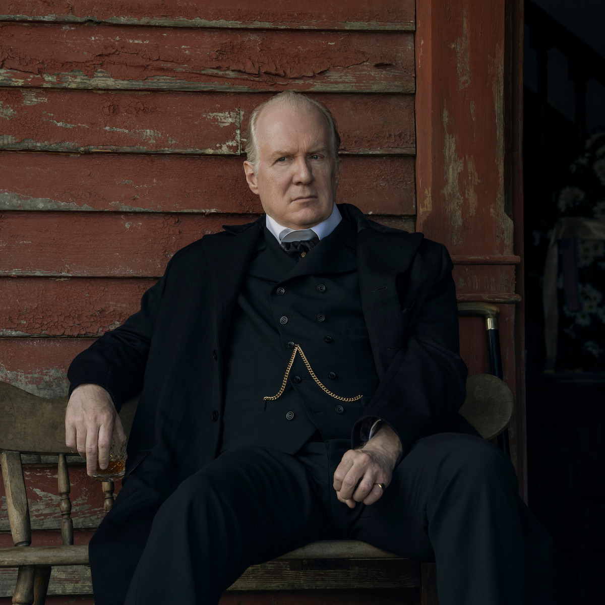 Tracy Letts como Cyrus Trask, de terno e casaco pretos, sentado em um banco rústico de madeira do lado de fora de uma construção desgastada de madeira vermelha, com um copo na mão, em um cenário de época.