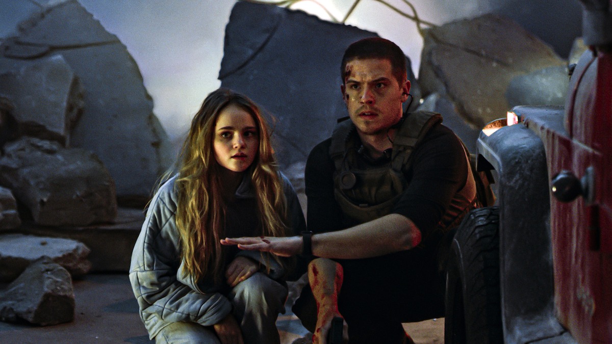 Megan Stott and Dylan Sprouse crouch next to each other amid cement debris in an image from the 2024 movie ‘Aftermath.’