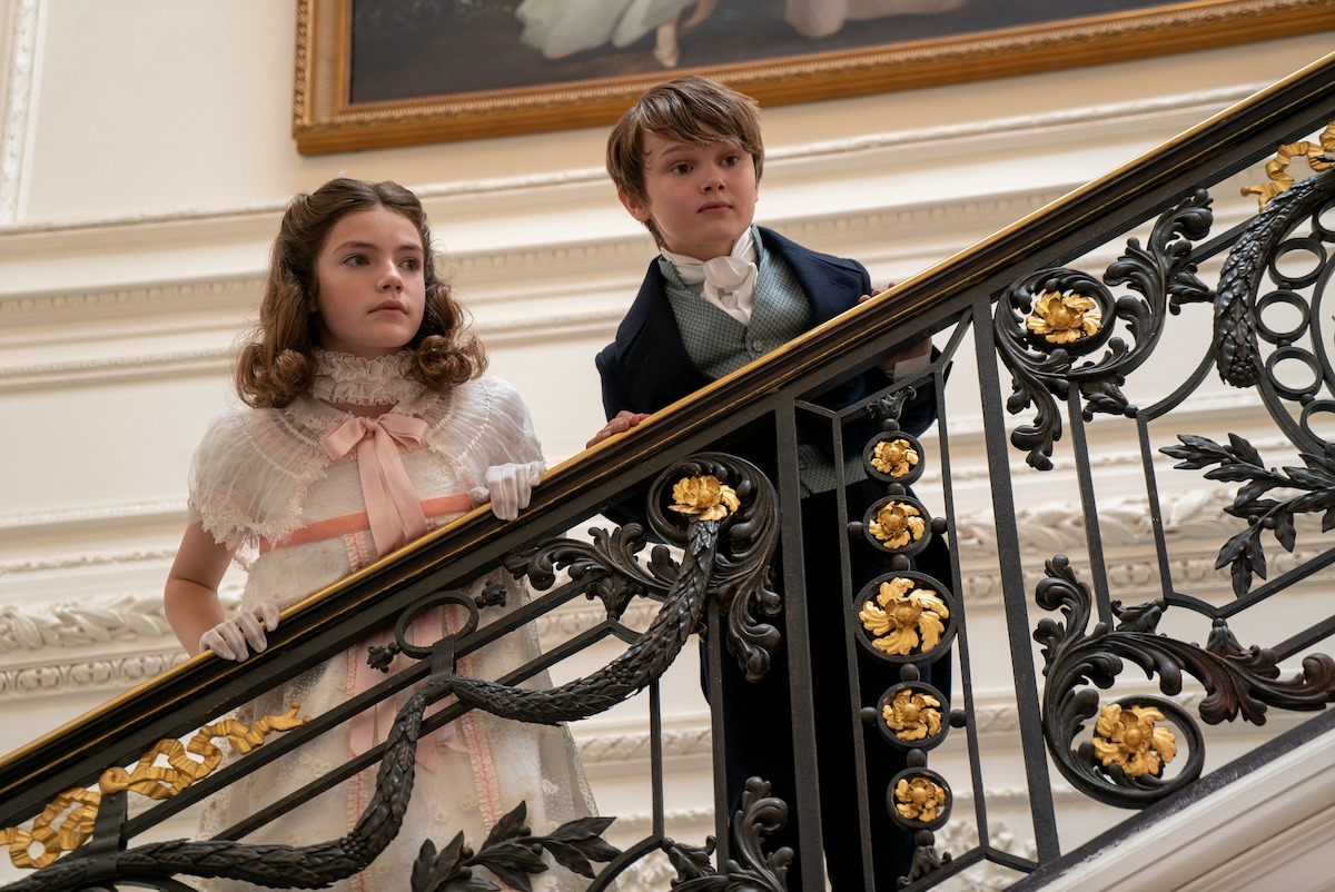 Two children in Victorian-era clothing stand on an ornate staircase with gold accents inside a lavish, historic building, gazing over the railing in a well-lit, elegant interior.