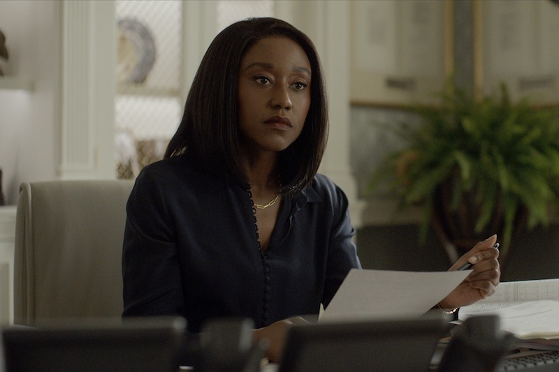 A woman with a serious expression sits at a desk holding papers and a pen, appearing focused and professional in an office setting.
