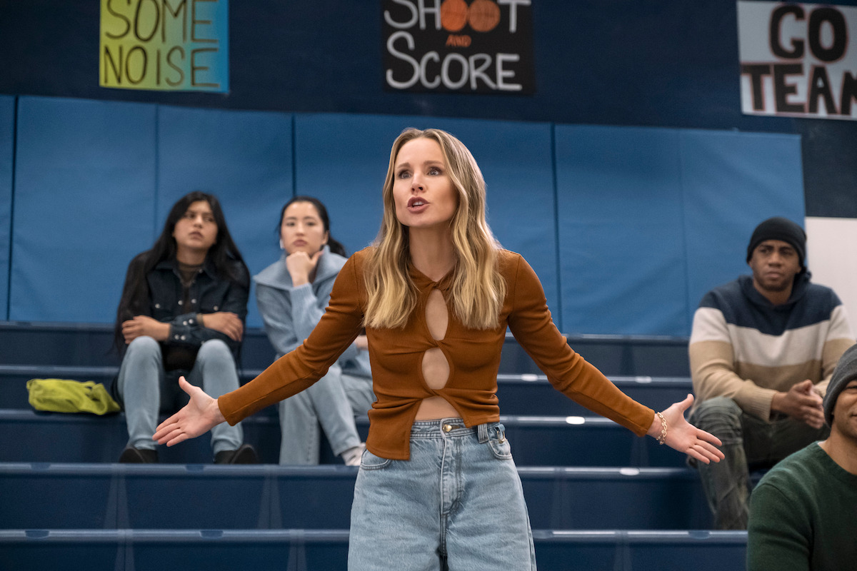Joanne (Kristen Bell) stands in the bleachers of a basketball court in jeans and a brown shirt.