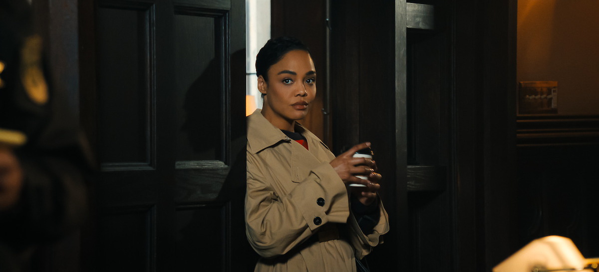 A woman in a beige trench coat holding a cup stands in a dark wooden doorway in a dimly lit room.
