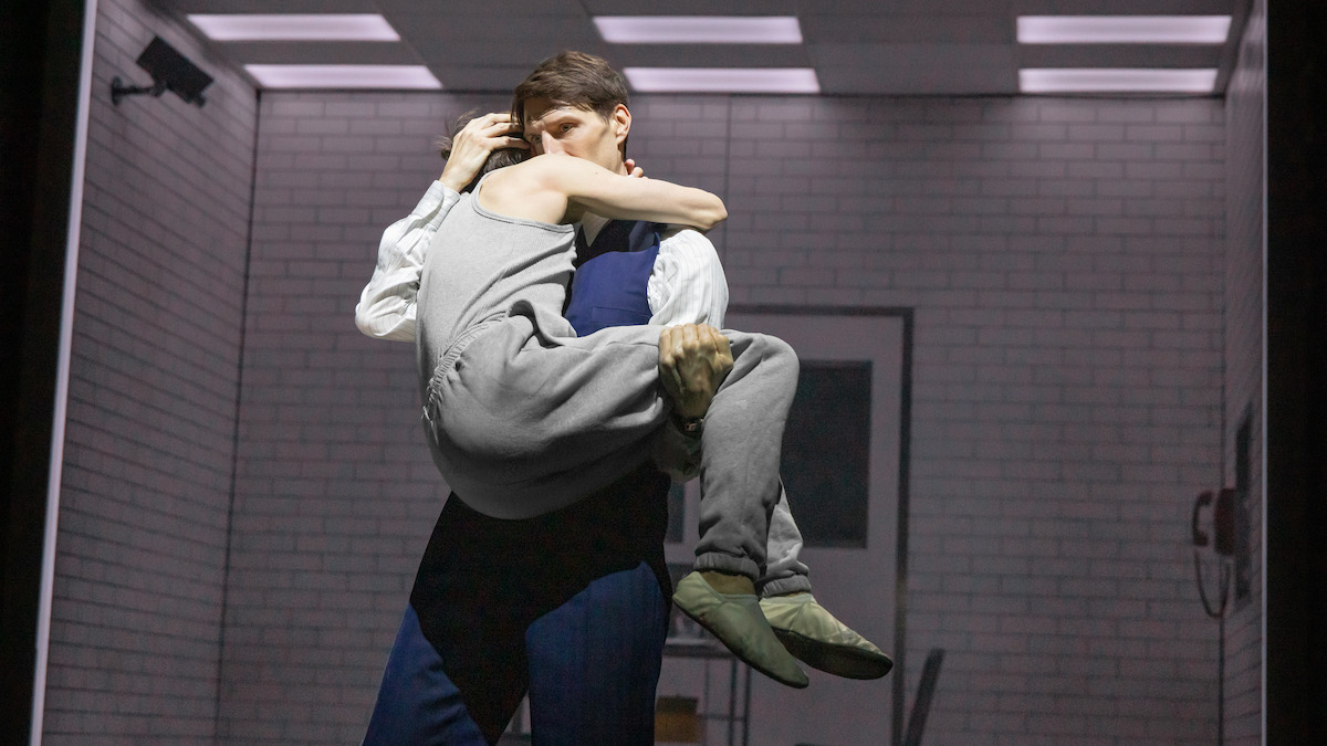 Two people embrace in a brightly lit sterile room with white brick walls, evoking a sense of intimacy and vulnerability in a clinical setting.