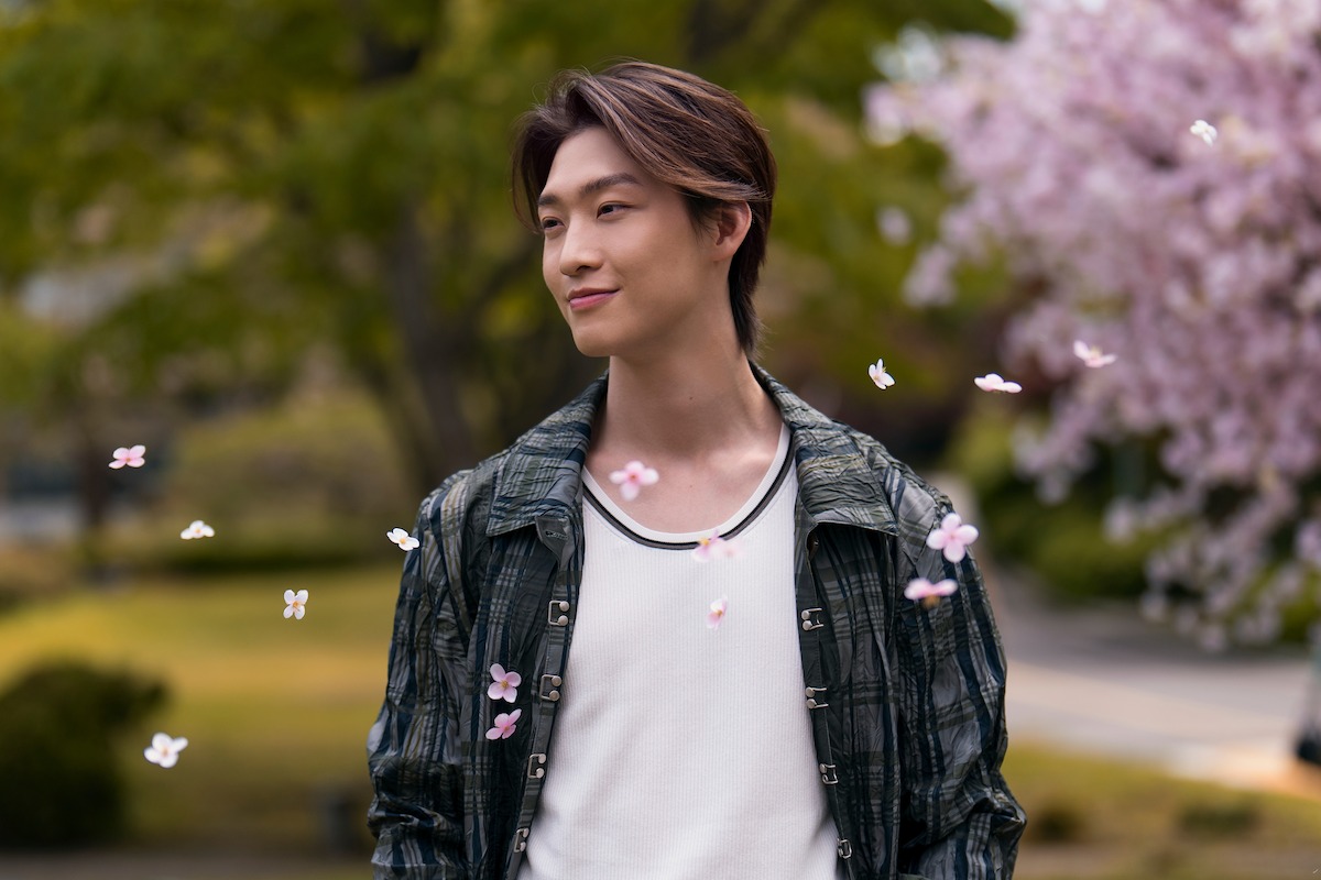 A young person stands outdoors among blooming cherry blossoms, smiling peacefully as petals fall around them; the background is a park-like setting with greenery and soft focus trees.