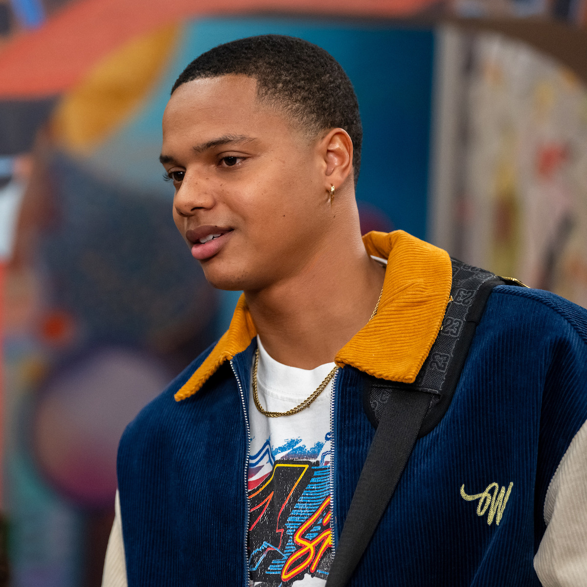 Elijah J. Roberts as Jalen with short hair and gold earring wearing a blue and yellow jacket stands indoors with colorful, blurred artwork in the background, looking slightly to the side and carrying a shoulder bag.