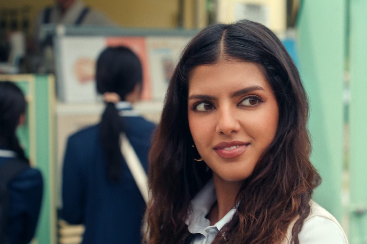 A woman with long dark hair and a white jacket stands outdoors, looking to the side with a slight smile. The background is out of focus, showing people in uniforms and pastel-colored structures, suggesting a public or social setting.