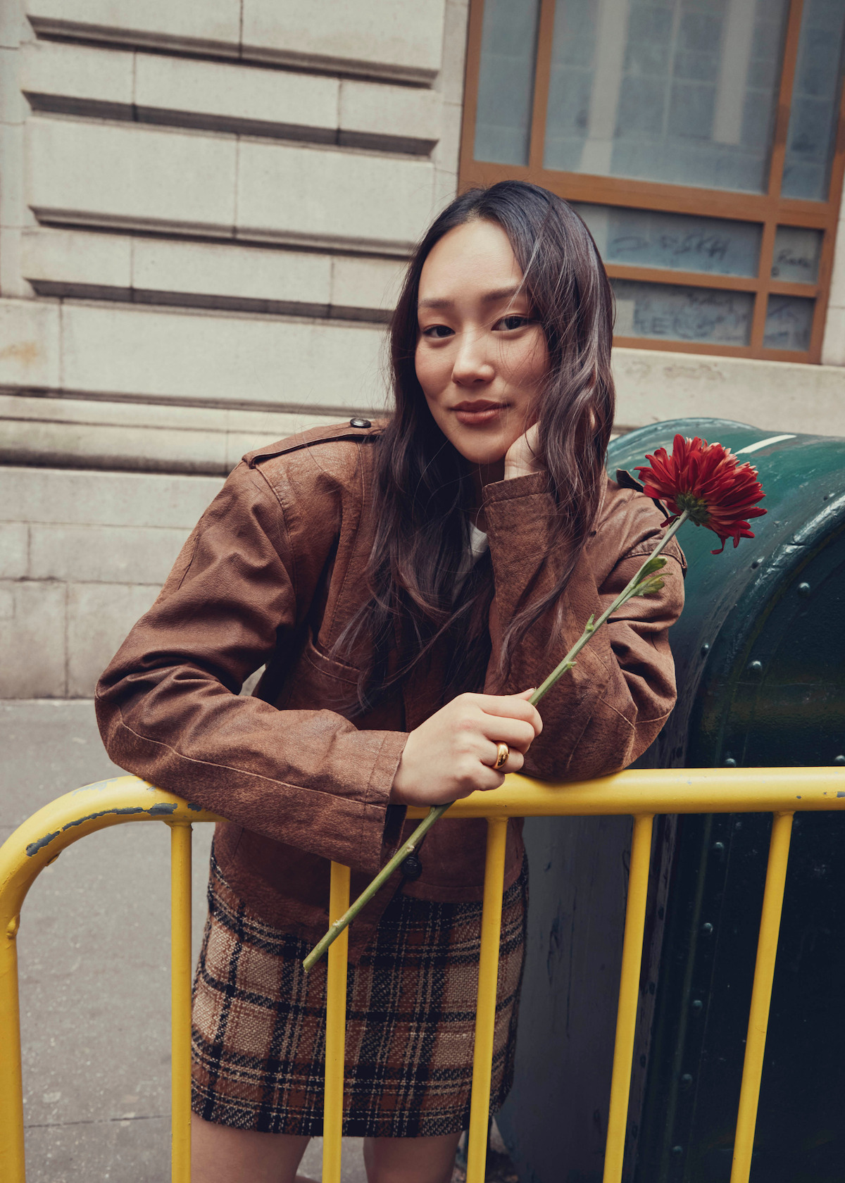 Woman with long dark hair holding a red flower, leaning on a yellow barrier in an urban environment with a stone building and green mailbox in the background.