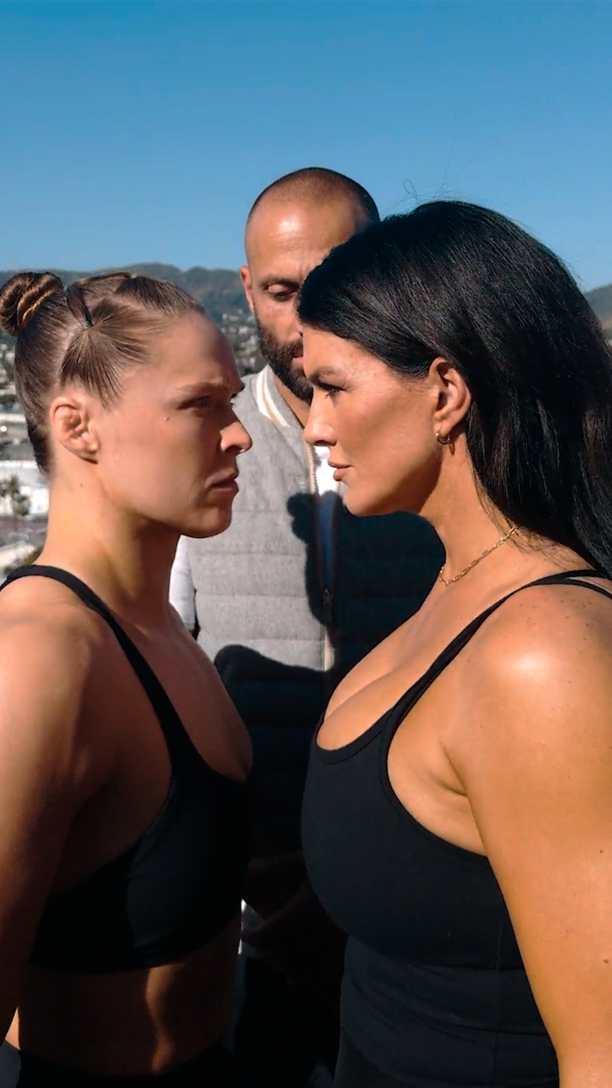 Two athletic women in sports bras face off intensely outdoors, with a man standing between them under a clear blue sky, mountains visible in the background, suggesting a pre-fight or competition setting.
