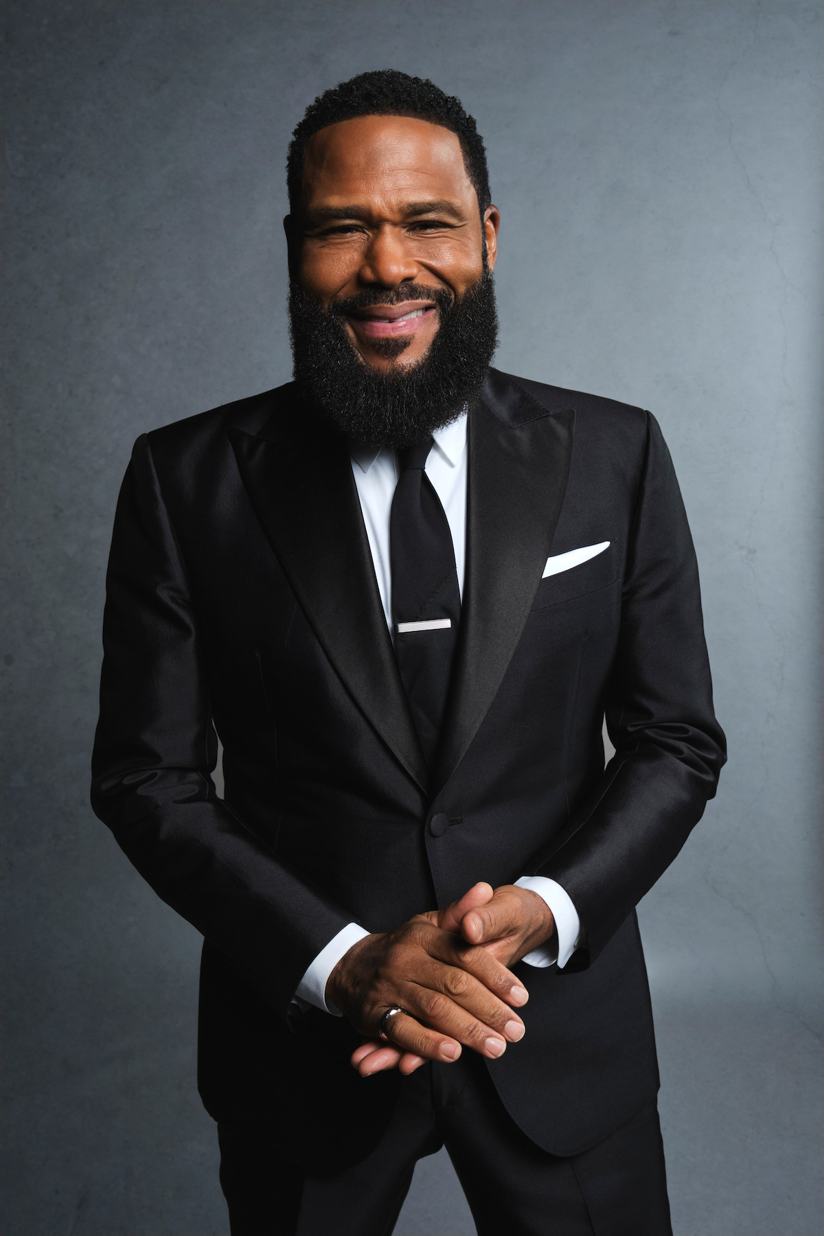 A confident man with a full beard smiles warmly while wearing a black tuxedo, white shirt, and tie. He stands against a neutral gray background, exuding elegance and a positive, approachable mood.