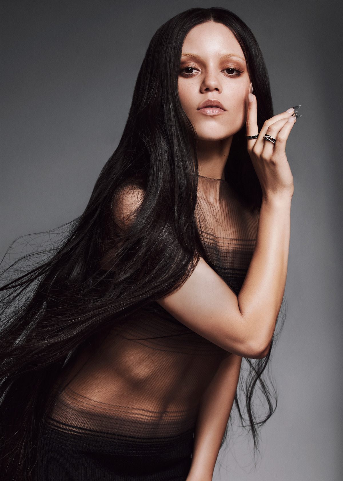 Jenna Ortega stands in front of a gray background looking to camera with a hand near her face wearing a layered sheer dress with long hair flowing down around her face and body.
