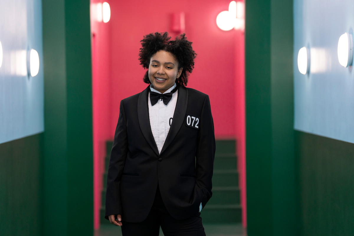 Person in a black suit and bow tie stands smiling in a brightly lit, colorful hallway with teal and pink walls, exuding a confident and cheerful mood.