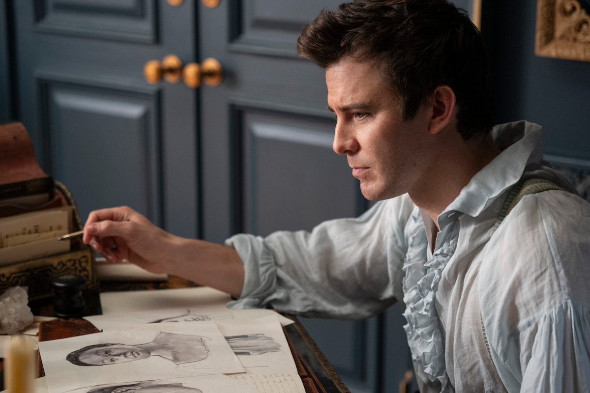 A man in historical clothing sits at a desk with sketches and drawing tools, appearing focused on his art in an elegant, dimly lit room with blue paneled walls and gold handles.