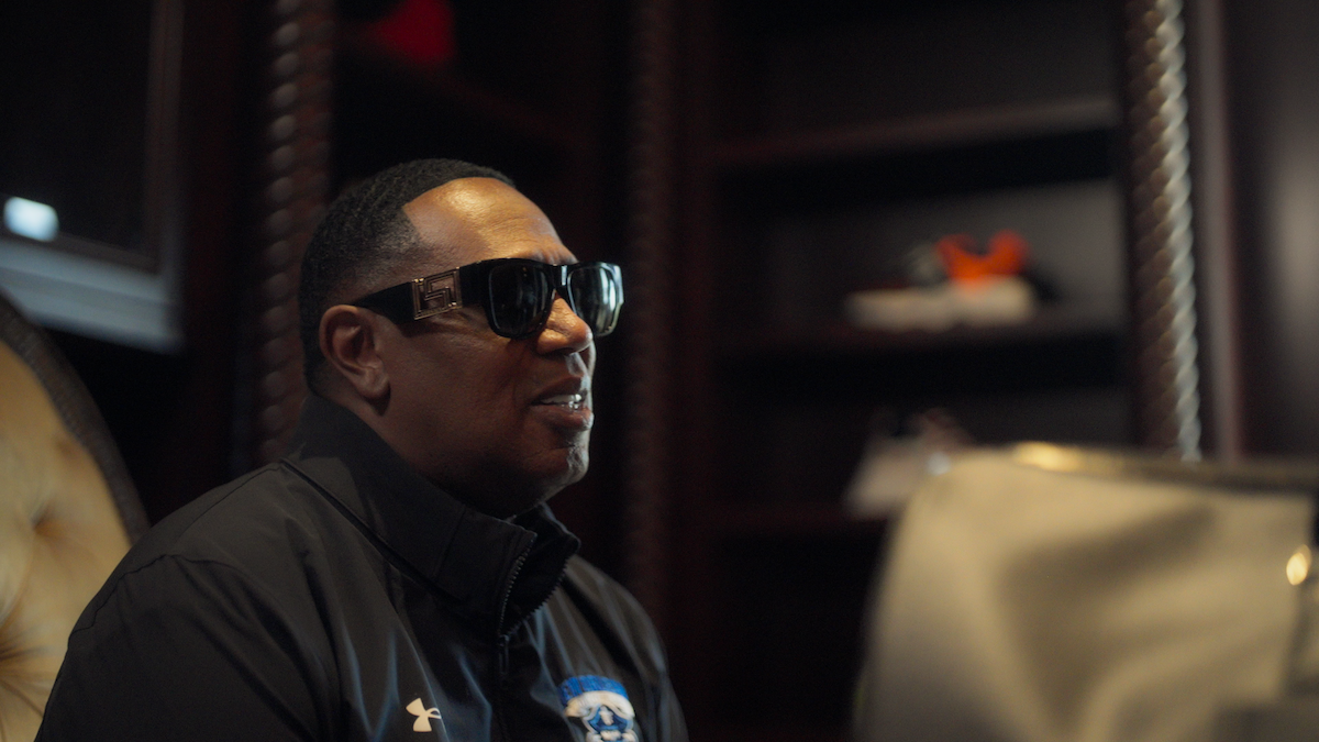 Man wearing sunglasses and a dark jacket sits in a dimly lit room with wooden shelves, a chair, and some shoes in the background. The mood is relaxed and the setting appears to be an office or study.