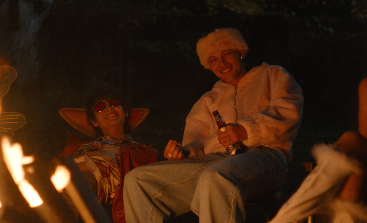 Two people in costumes sit by a campfire at night, smiling and enjoying a relaxed outdoor setting; one holds a drink and both wear warm, playful outfits.