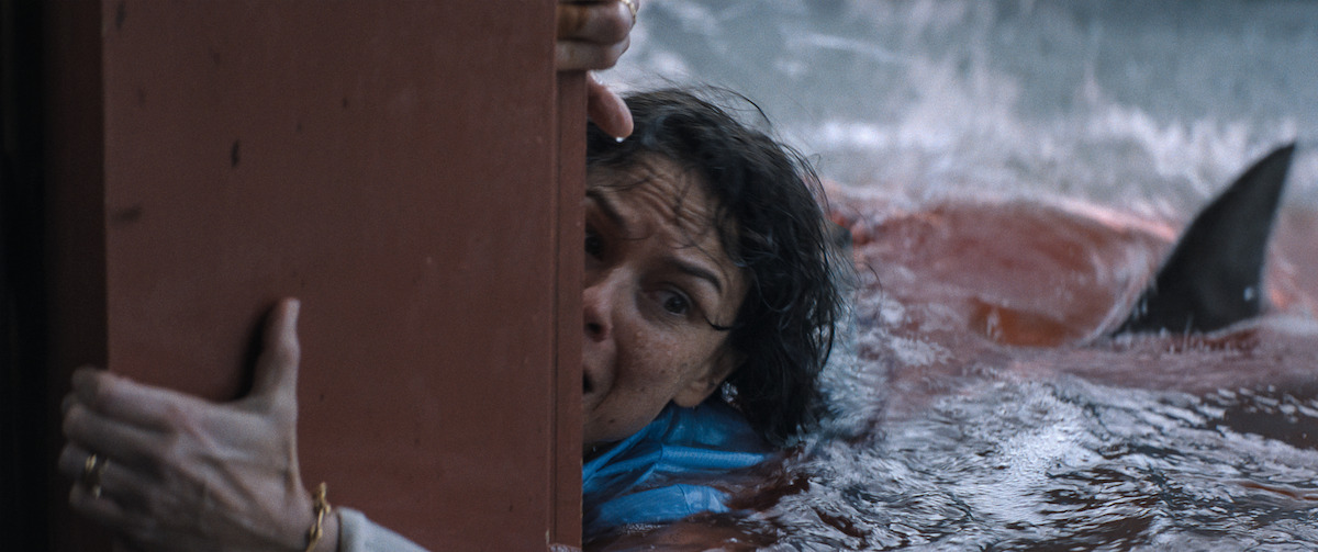 A terrified woman clings to a red door in rough, bloody water as a shark fin approaches behind her, suggesting a dangerous and intense aquatic environment.