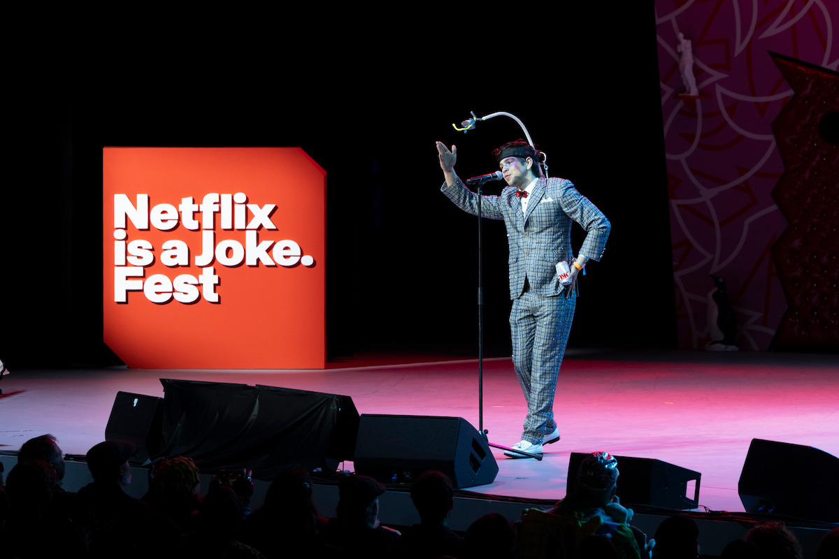 A performer in a suit, clown makeup, and bowler hat entertains an audience on stage at the Netflix is a Joke Fest, with a large red event sign and playful stage décor in the background.