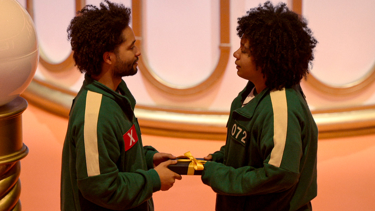 Two people in green uniforms exchange a gift-wrapped box in a warmly lit, stylized room, creating a tense but intimate atmosphere.