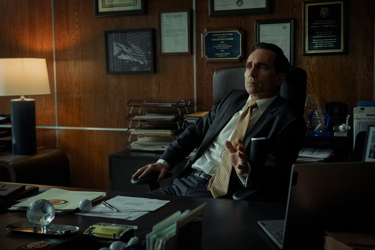 A man in a suit sits at a desk in a dimly lit office with wood-paneled walls, paperwork, and award plaques behind him, looking serious and gesturing with his hand.