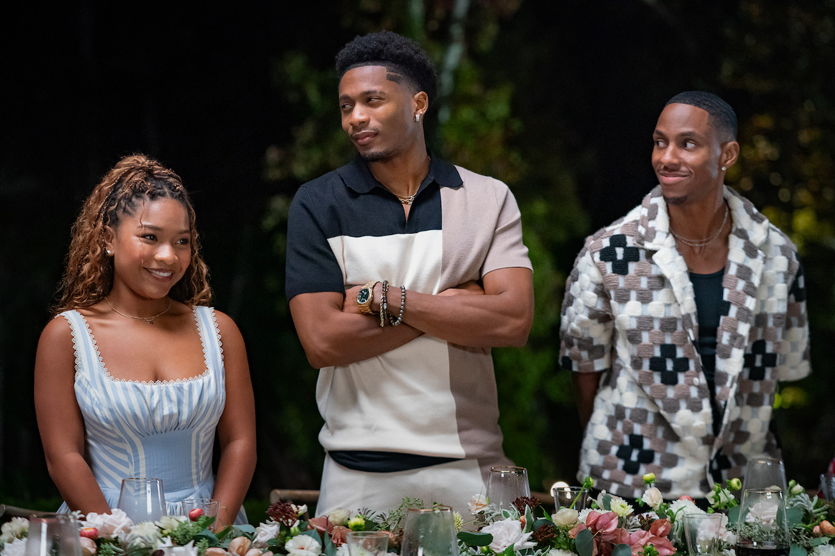 Three people standing behind a decorated dinner table outdoors at night, dressed in stylish casual attire, surrounded by flowers and candles, appearing to enjoy a social gathering or celebration.