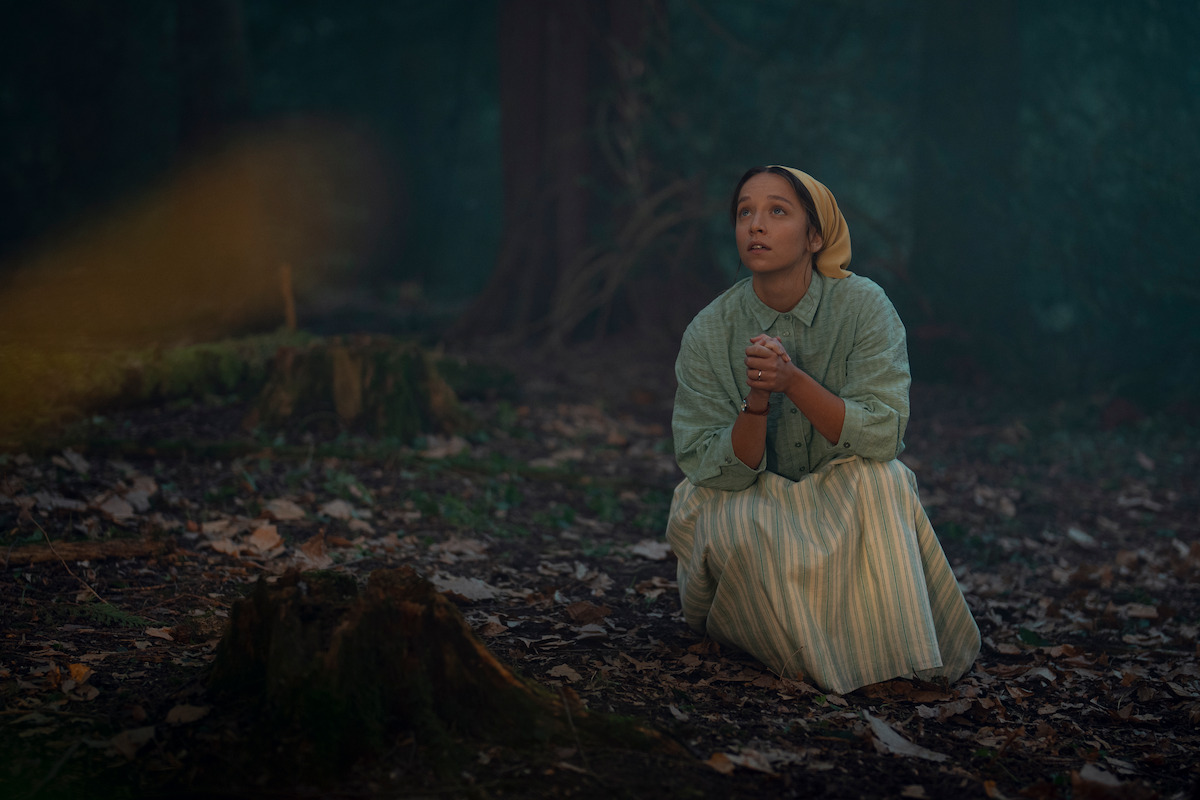 Woman in old-fashioned clothing kneels on forest floor among tree stumps and fallen leaves, looking upward with clasped hands; the atmosphere is dim and misty, suggesting a historical or dramatic setting.