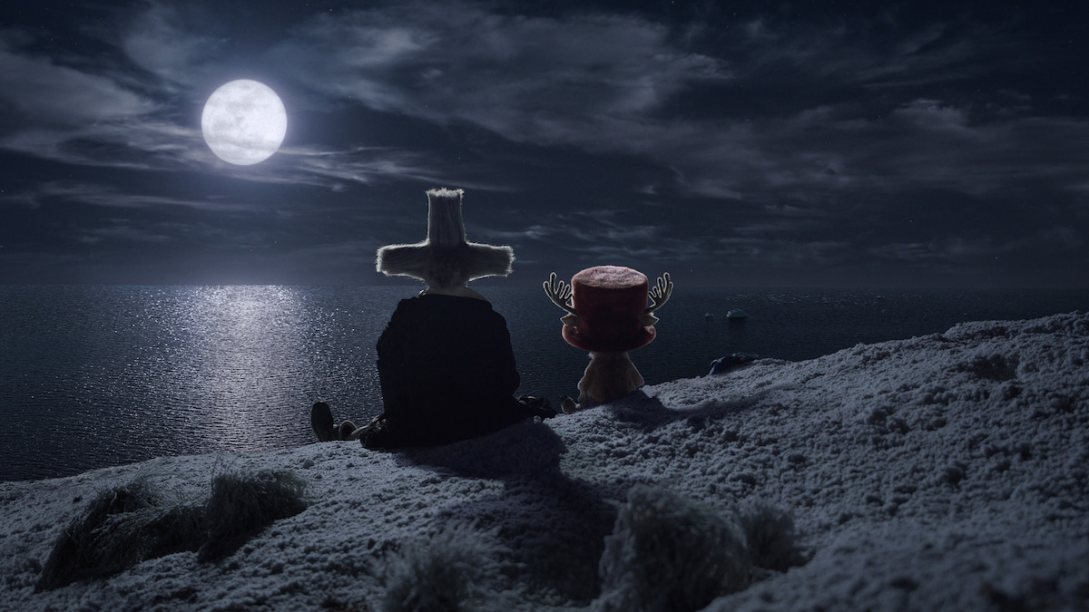 Mark Harelik as Dr. Hiriluk and Tony Tony Chopper sit on a snowy hill overlooking a moonlit sea at night, with clouds in the sky and the full moon reflecting on the water, creating a peaceful and contemplative atmosphere.