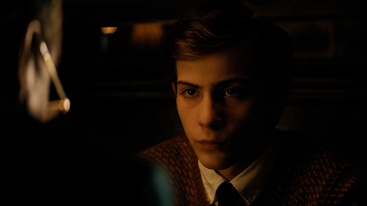 A young man in a brown sweater and tie sits in a dimly lit room, staring intently at someone across a table. The lighting is low and warm, suggesting an intimate or serious conversation in a quiet, possibly vintage setting.