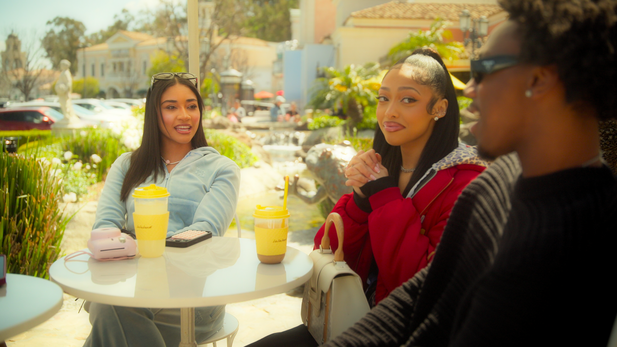 Three friends sit at an outdoor café table with drinks, chatting on a sunny day. The background shows greenery, water, and elegant buildings, creating a relaxed, social atmosphere.
