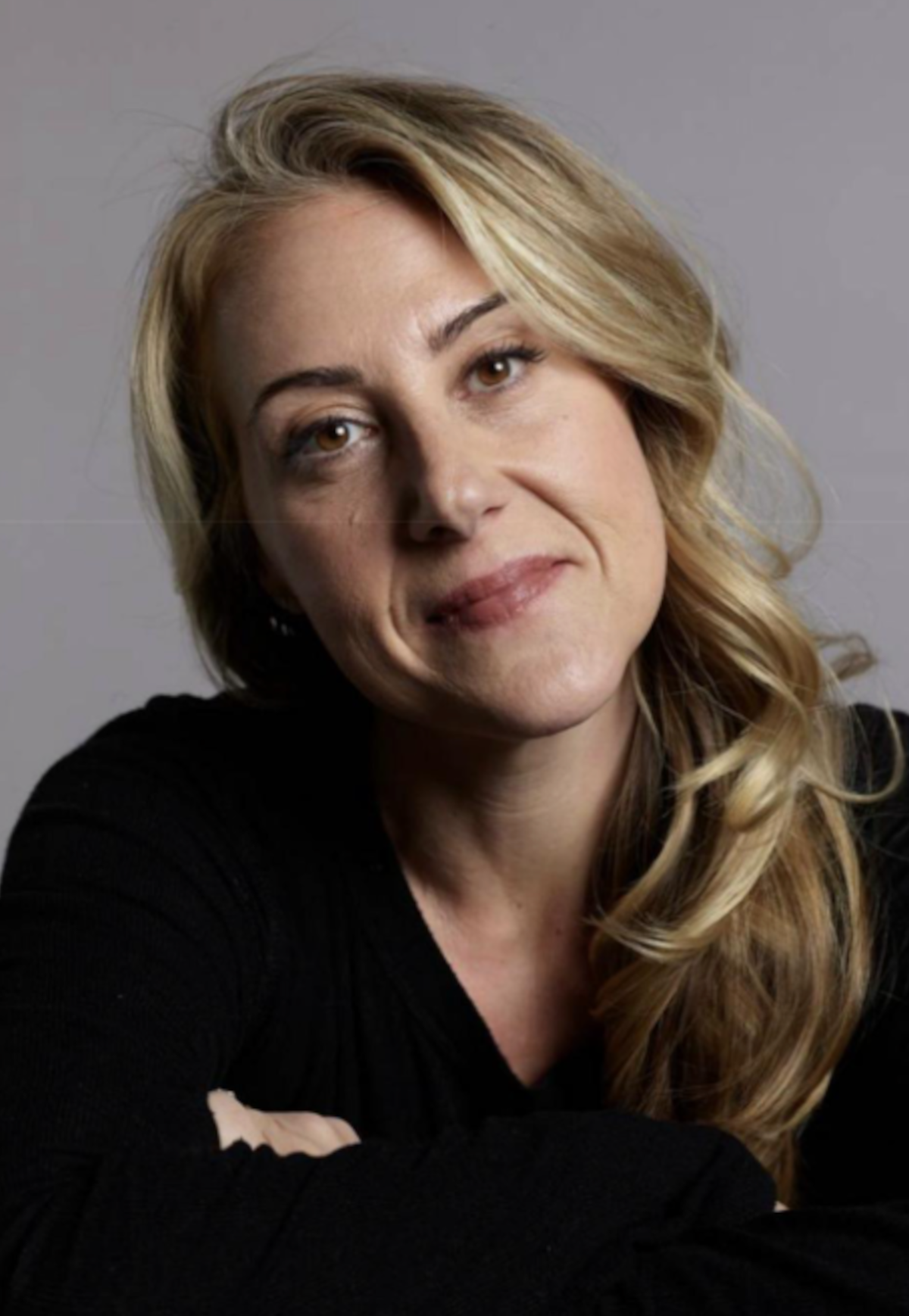 Woman with long blonde hair wearing a black top, sitting indoors against a plain gray background, looking at the camera with a slight smile and relaxed posture.