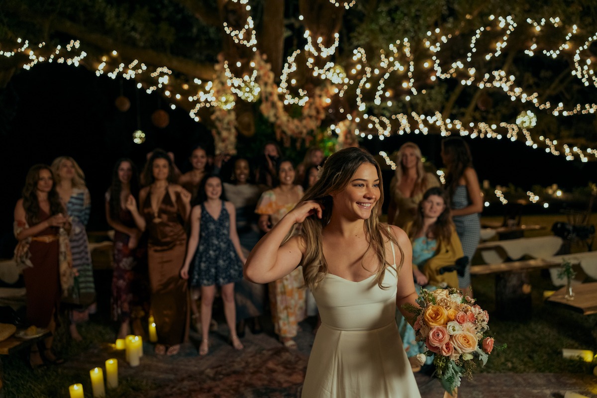 A bride holding a bouquet stands smiling outdoors at night, preparing to toss flowers to a group of women behind her under a tree decorated with string lights and surrounded by candles.