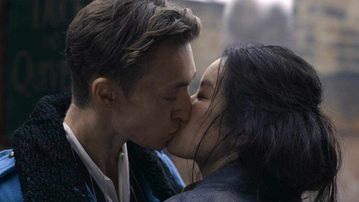 Two people share a passionate kiss outdoors, dressed in coats, with a blurred urban background suggesting a city or street setting.