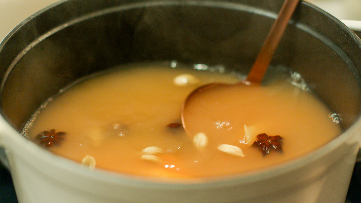A steaming pot of spiced orange liquid, with visible star anise, nuts, and a ladle, suggesting a cozy kitchen environment and a warm, comforting mood.