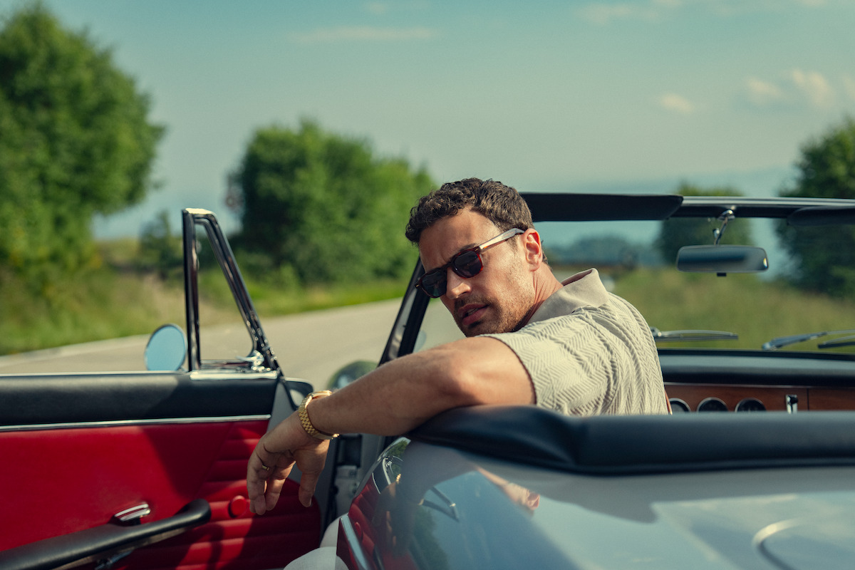 Man wearing sunglasses sits in a classic convertible car with the door open on a country road, surrounded by greenery and trees under a clear sky, creating a relaxed and stylish atmosphere.