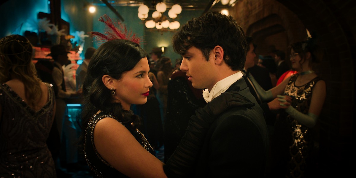 A woman and man in vintage formal attire dance closely at a lively, dimly lit party with guests dressed in 1920s-style clothing, surrounded by warm lighting and festive decorations.