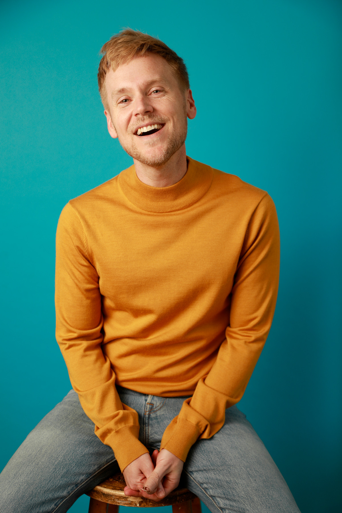 Smiling man with light hair wearing a mustard yellow sweater and jeans, sitting on a stool against a plain teal background in a studio setting.