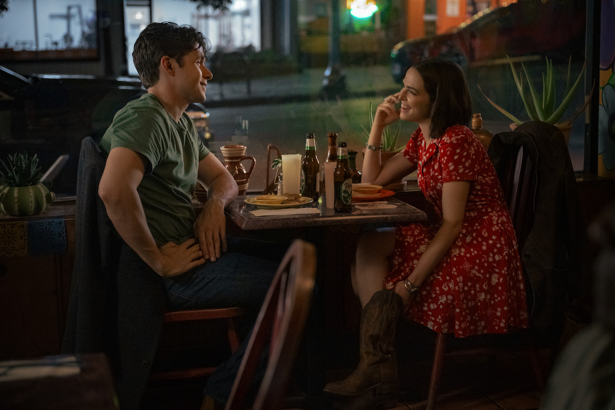 A man and woman sit across from each other at a cozy restaurant table at night, smiling and talking over food and drinks, with a street visible through the window behind them.