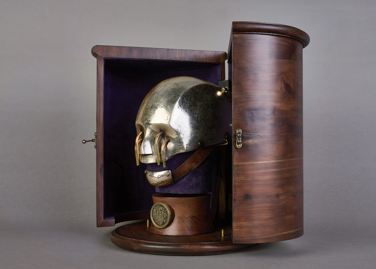 A curved wood box holds a silver helmet in the shape of a human head being held up by a purple velvet stand displayed against a gray background.