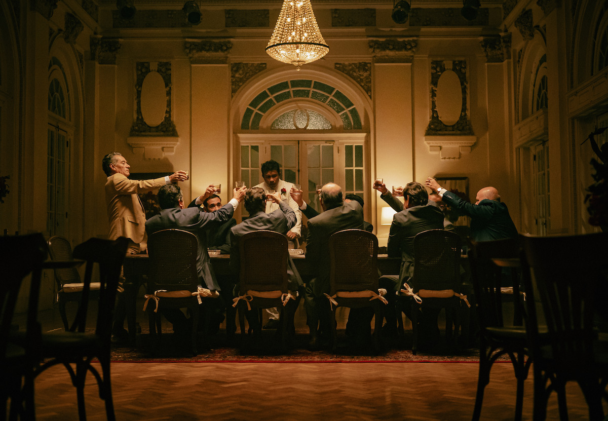 Homens em volta de uma mesa fazem um brinde em cena de “Os Donos do Jogoä