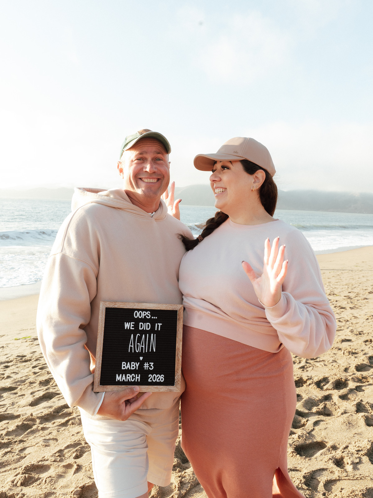 Smiling couple on a sandy beach, dressed in casual clothes and hats, holding a sign announcing their third baby due in March 2026, with ocean and distant hills in the background on a bright day.