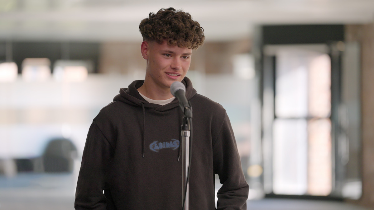 Young man with curly hair wearing a dark hoodie stands in front of a microphone, indoors with soft daylight and blurred background, giving a relaxed and slightly nervous vibe.