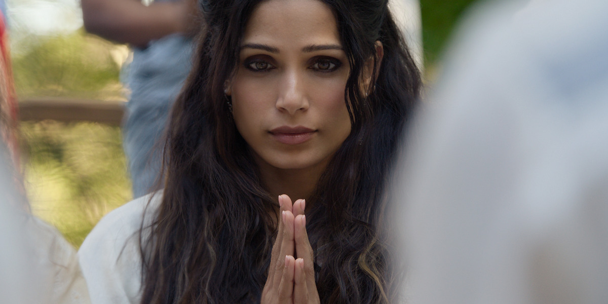 Woman with long dark hair sitting outdoors, hands pressed together in a prayer gesture, looking ahead with a calm, serene expression, surrounded by blurred figures in a natural, sunlit environment.