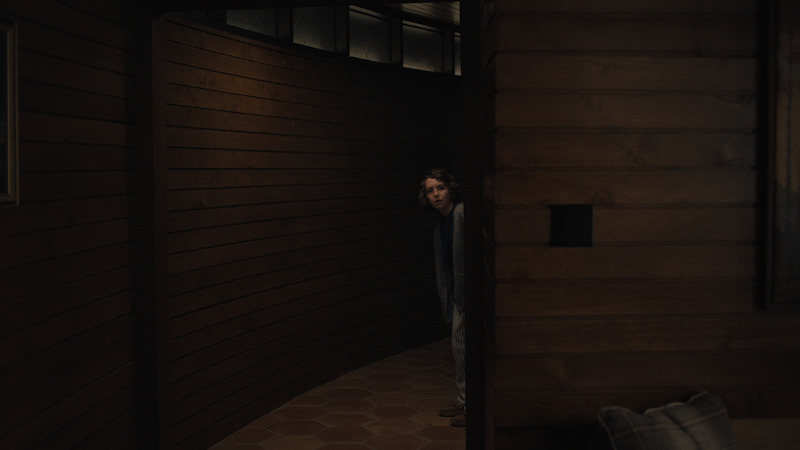 A person cautiously peeks around a curved wooden hallway in a dimly lit, cozy interior featuring wood-paneled walls, tile floor, and a partially visible couch with cushions in the foreground.