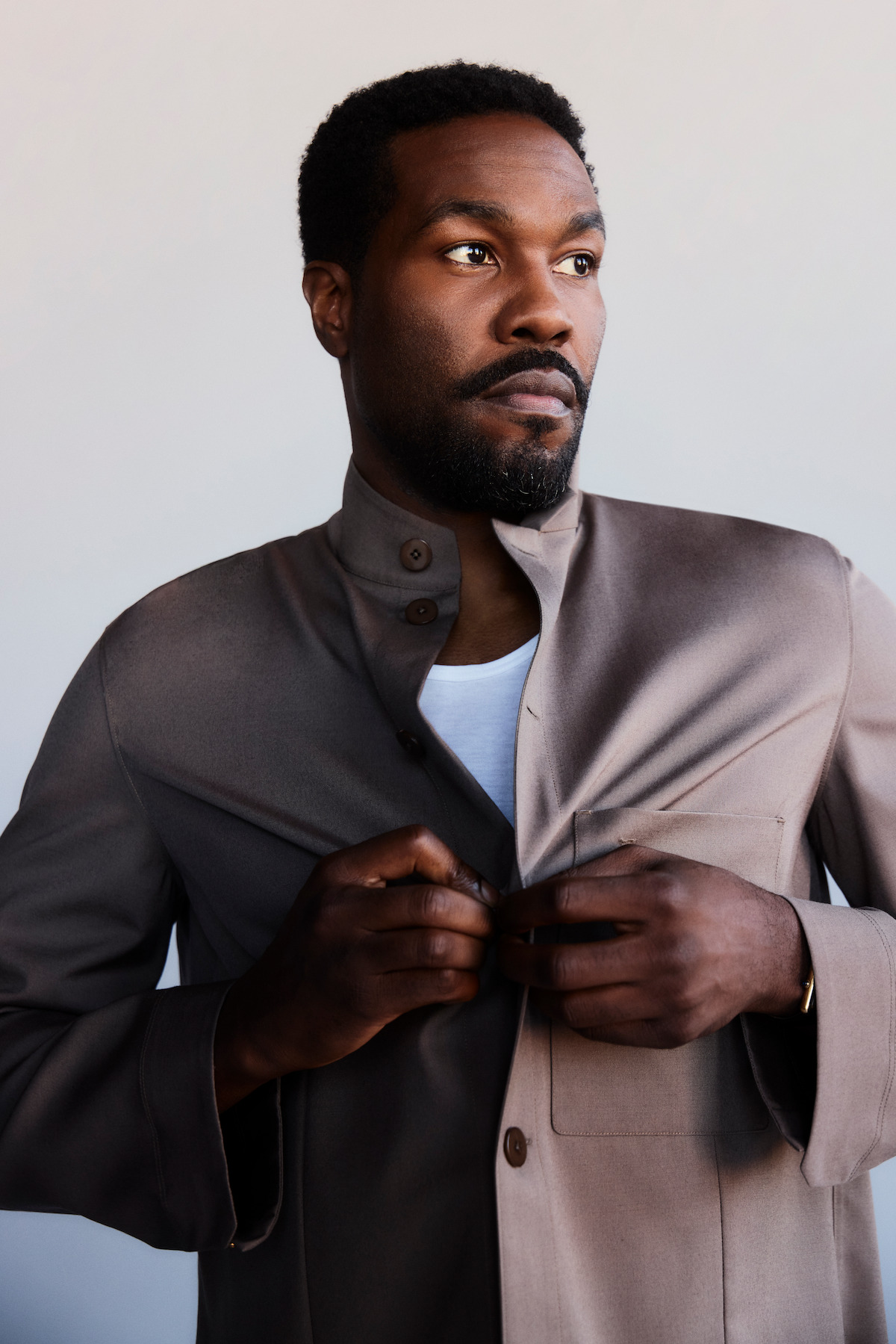 Man with short hair and beard wearing a taupe jacket over a white shirt, standing indoors against a plain light background, looking thoughtfully to the side while adjusting his jacket.