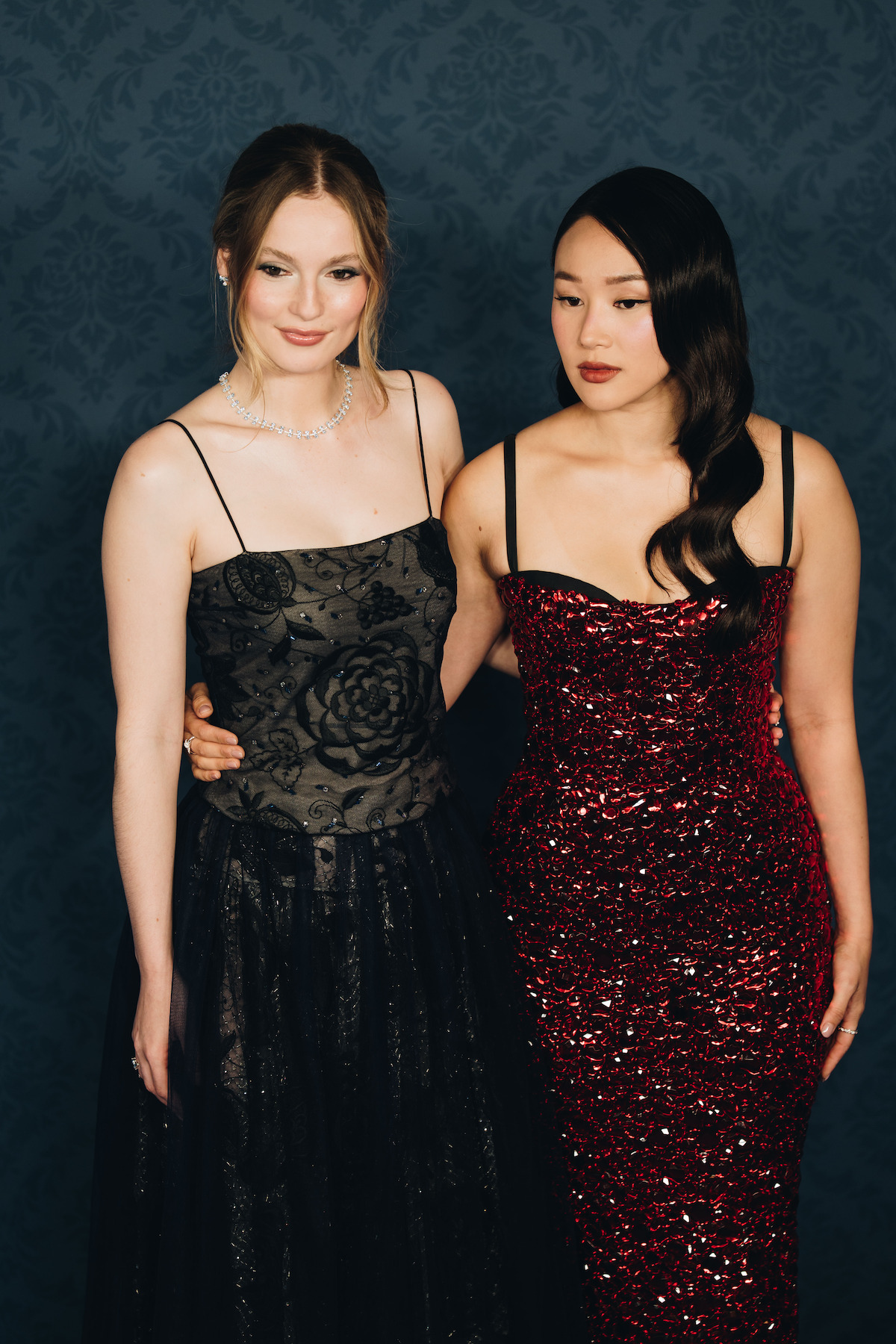 Duas mulheres com roupas formais, uma com um vestido preto estampado com flores e a outra com um vestido vermelho de paetês, estão posando lado a lado para uma foto. Elas estão diante de um painel escuro e estampado em um evento elegante.